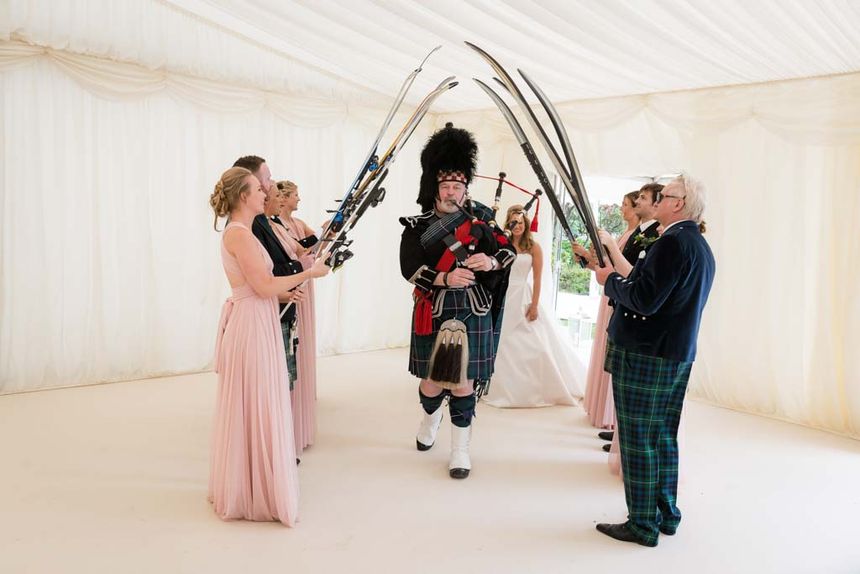 Bridesmaids and groosmen lined up while holding fishing rods for the wedding procession, and a man wearing a plaid while playing a flute is walking